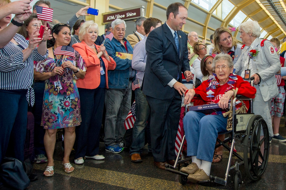 People wave small American flags as a veteran in a wheelchair proceeds past hem in an airport hall.