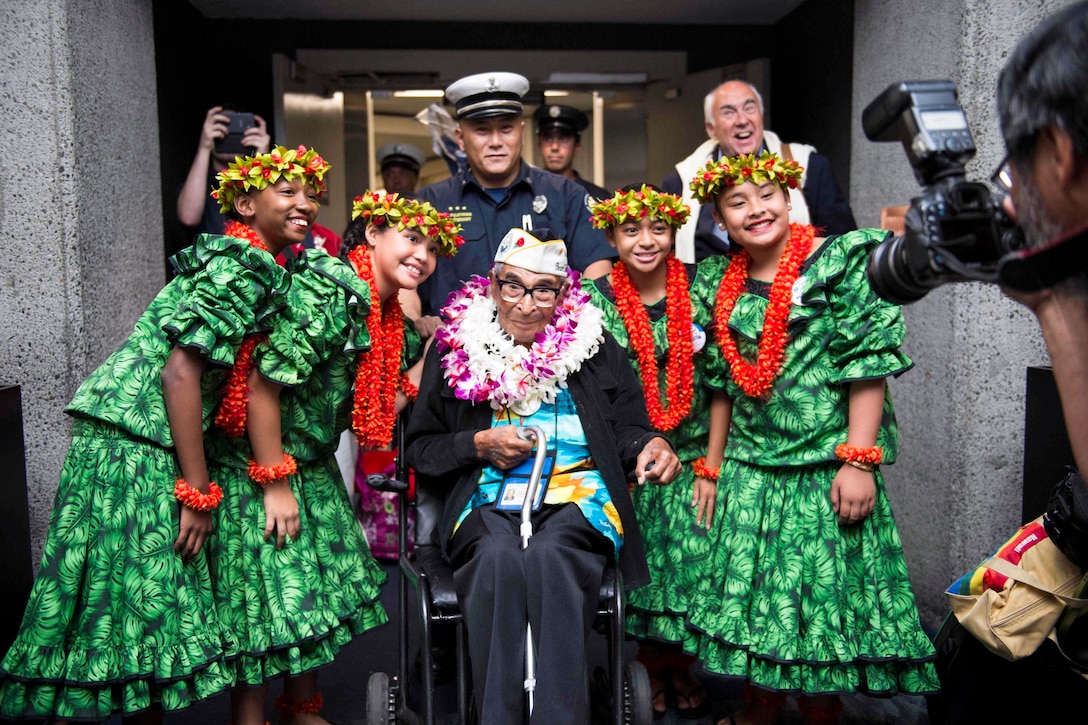 A veteran in a wheelchair flanked by smiling hula dancers smiles for a photo.