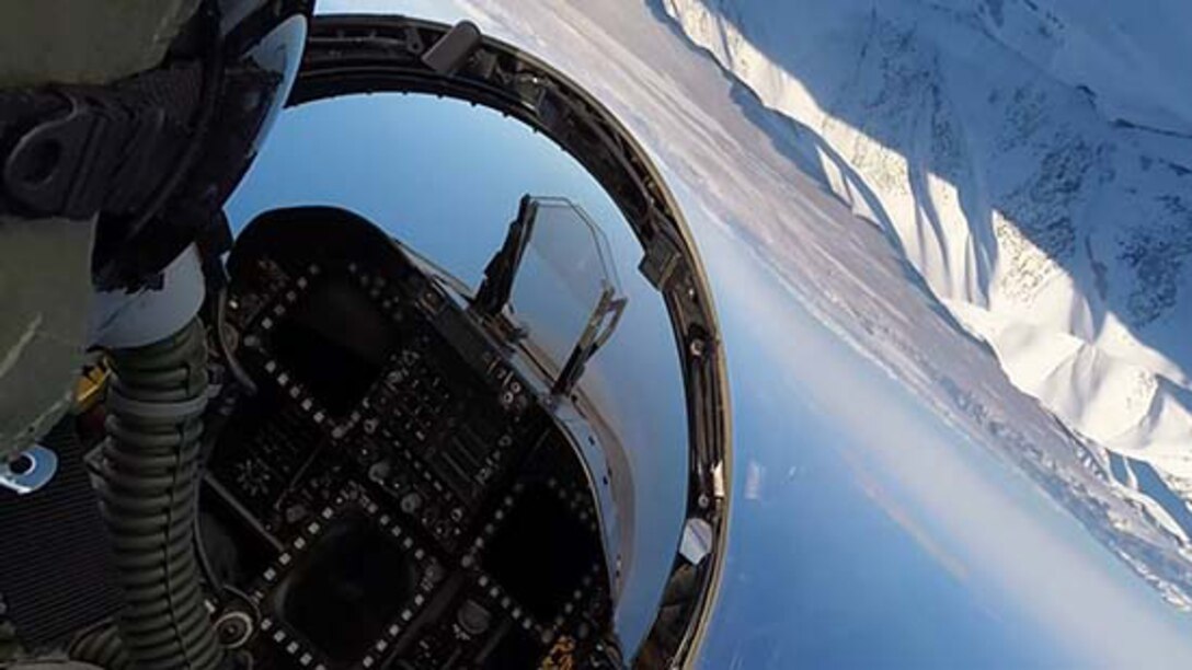 An over-the-shoulder view of a pilot in a cockpit performing a maneuver nearly upside-down with mountains in the background