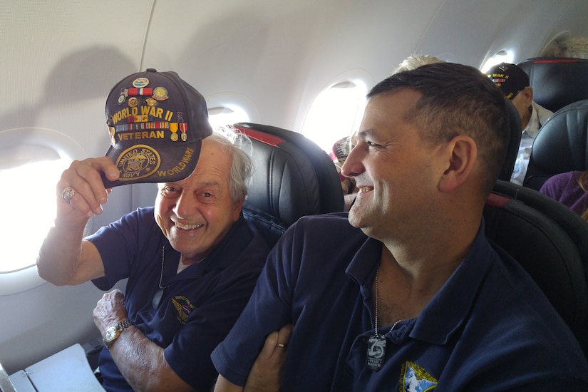 A World War II veteran tips his cap toward the camera while in an airplane window seat.