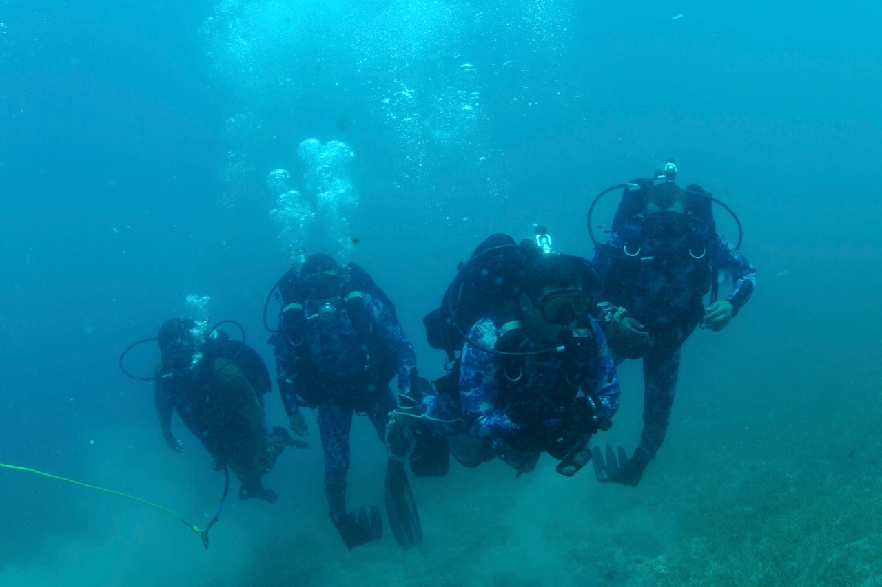 Four sailors swim underwater in scuba diving gear.