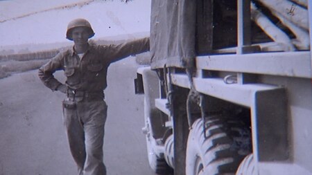 Black and white photo with soldier leaning up against a old Army truck.