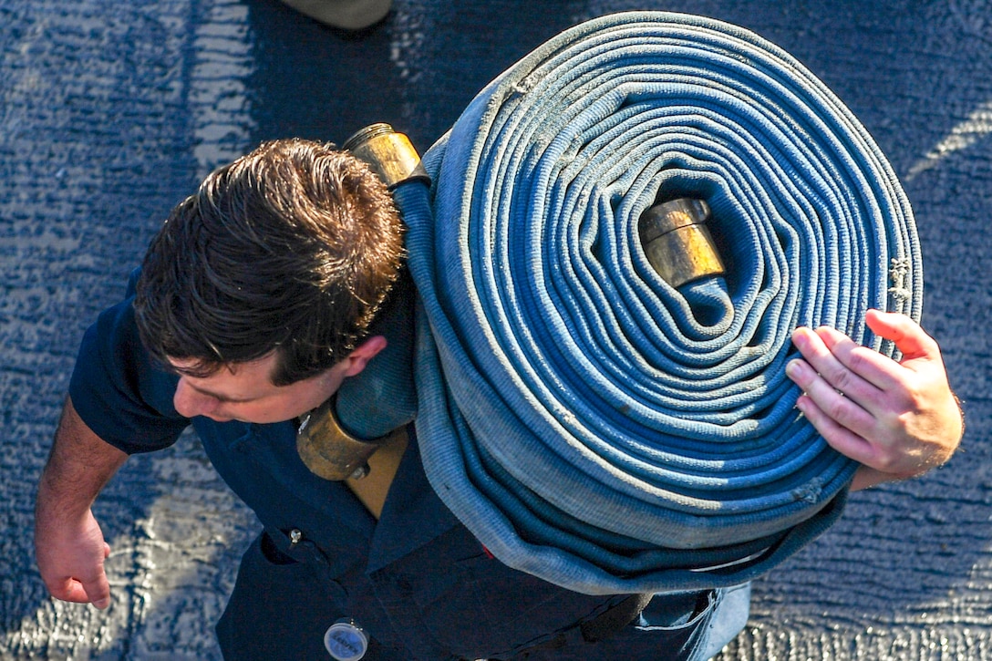 A sailor carries a coiled hose on his shoulder