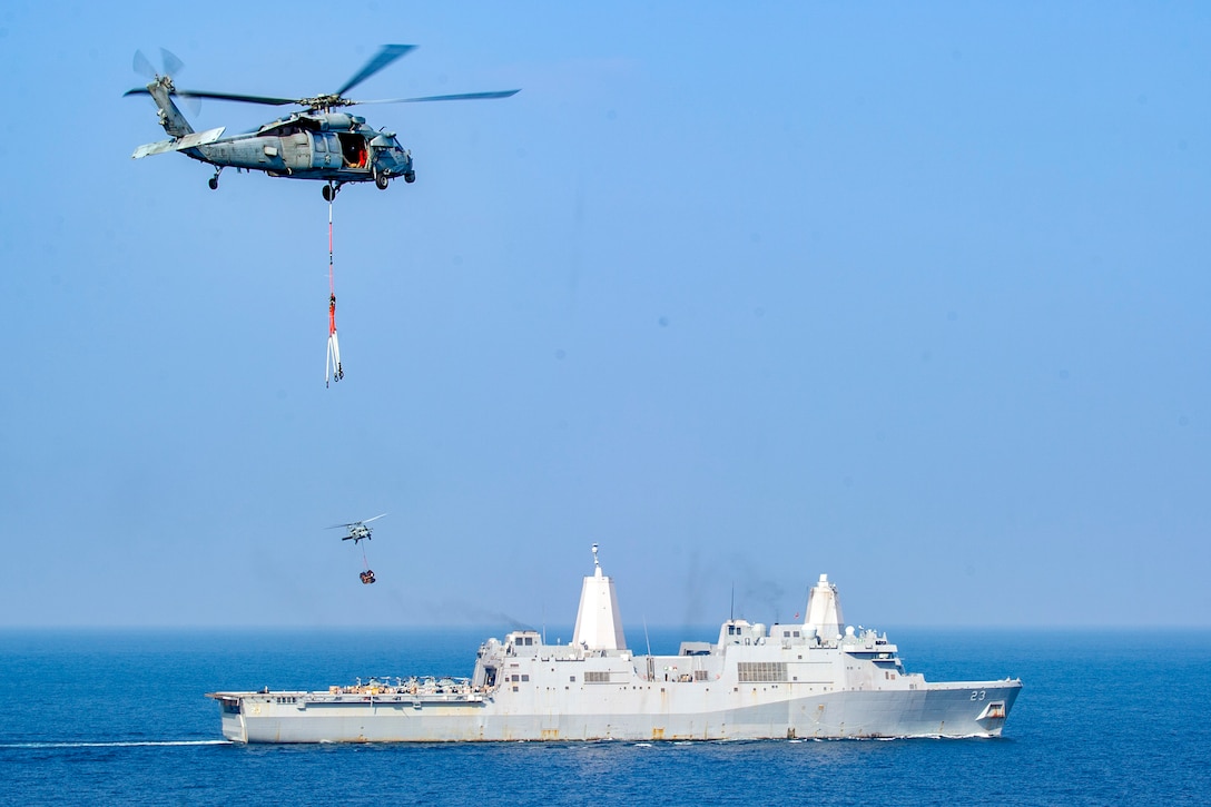 A helicopter lowers supplies onto a naval vessel at sea.