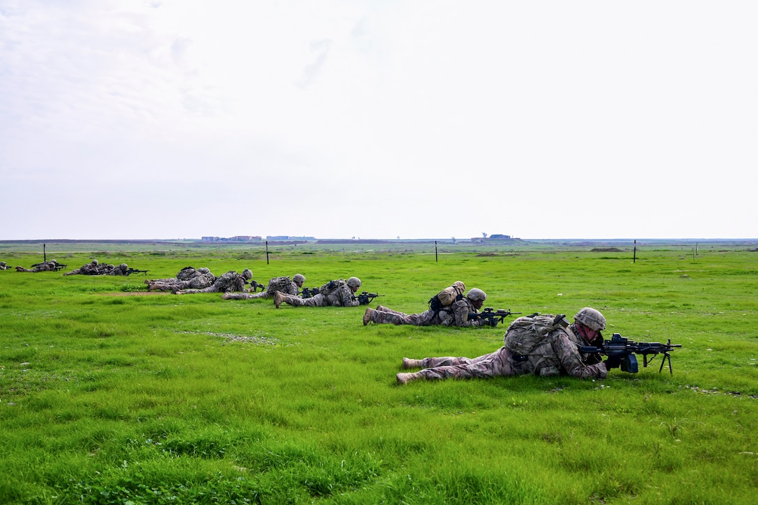 A group of soldiers lie down on the ground in  a row pointing their weapons.