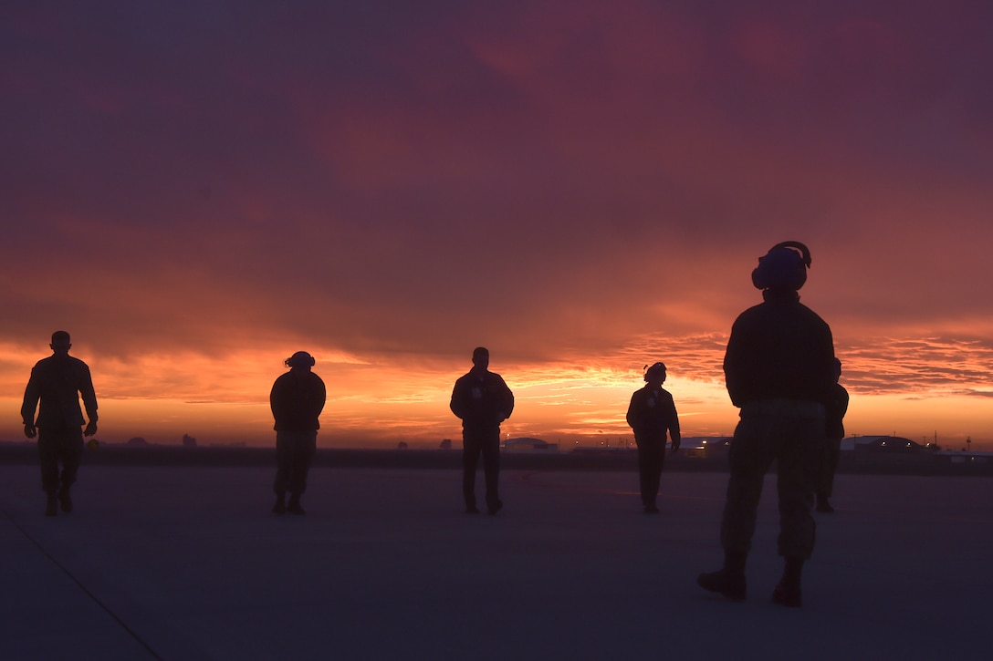 Sailors, shown in silhouette, walk on a flightline under a dark pink- and blue-streaked sky.