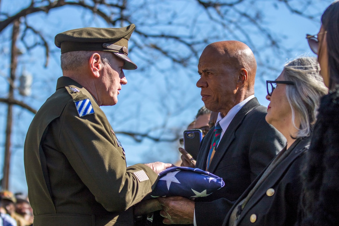 A soldier hands a folder U.S. flag to civilian in  a suit.