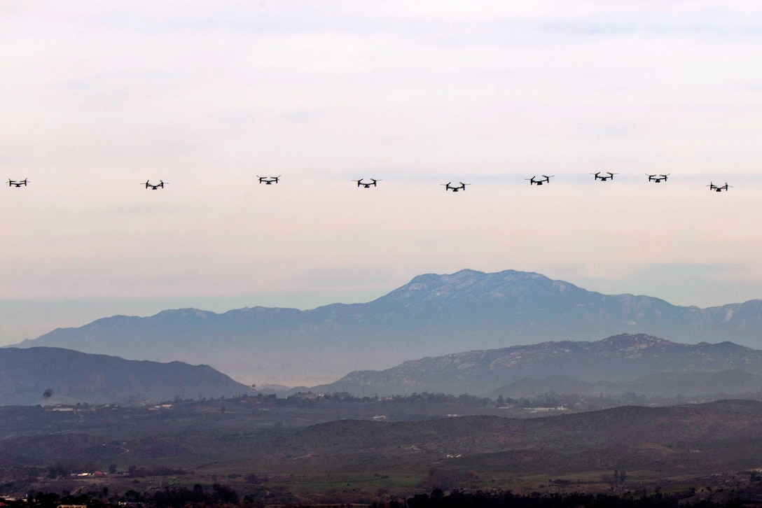 Nine tiltrotor aircraft fly in a horizontal row over a mountain range.