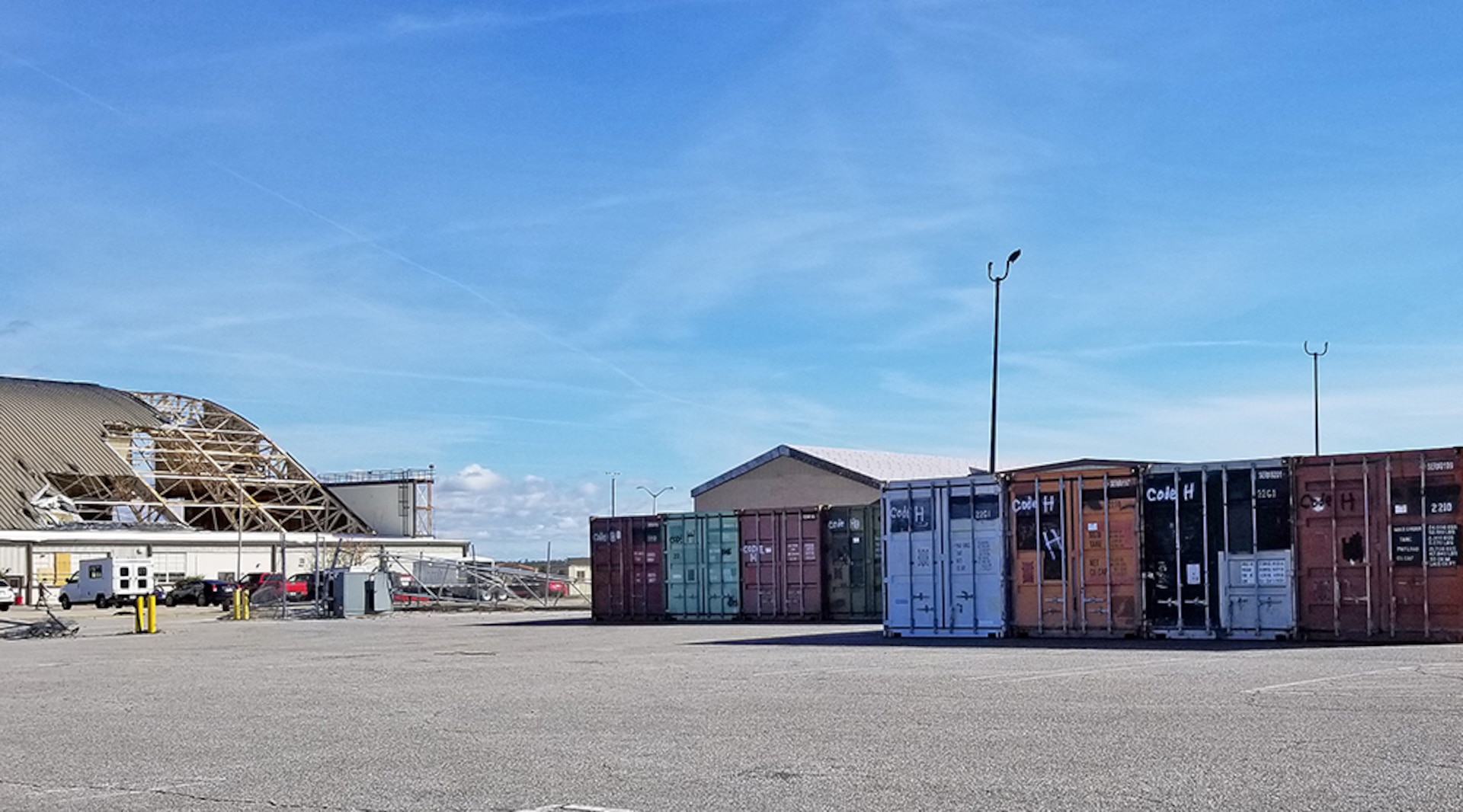 Excess shipping containers supplied from DLA Disposition Services line the parking lot of a damaged hangar facility at Tyndall AFB.