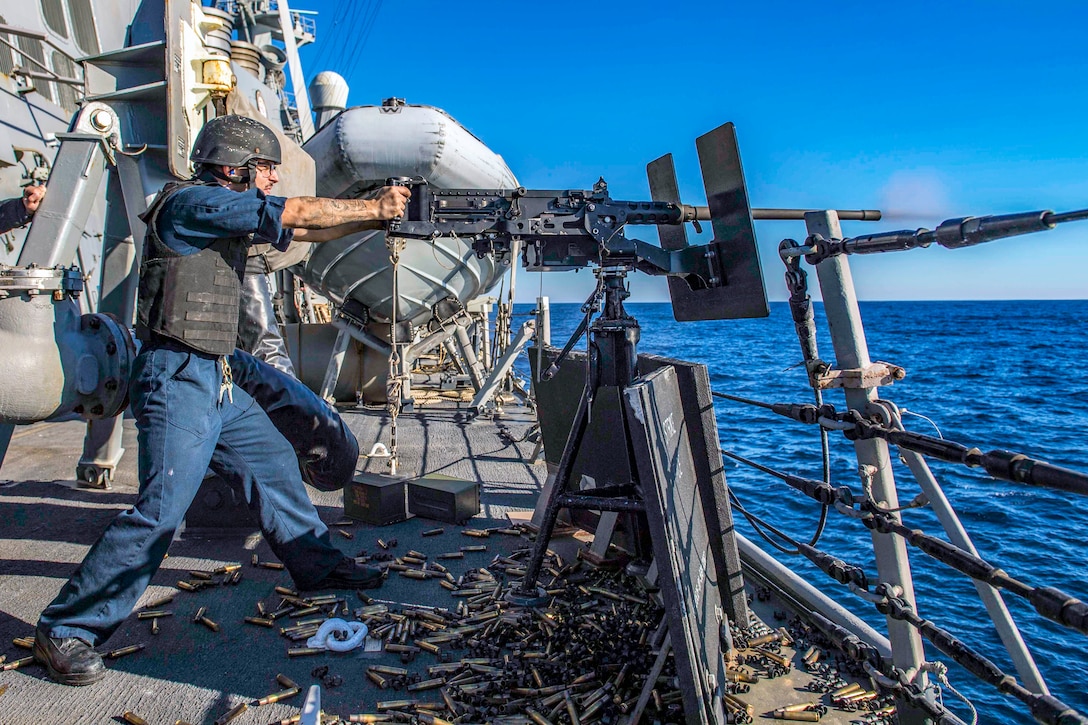 A sailor fires a big gun off the bow of a ship.