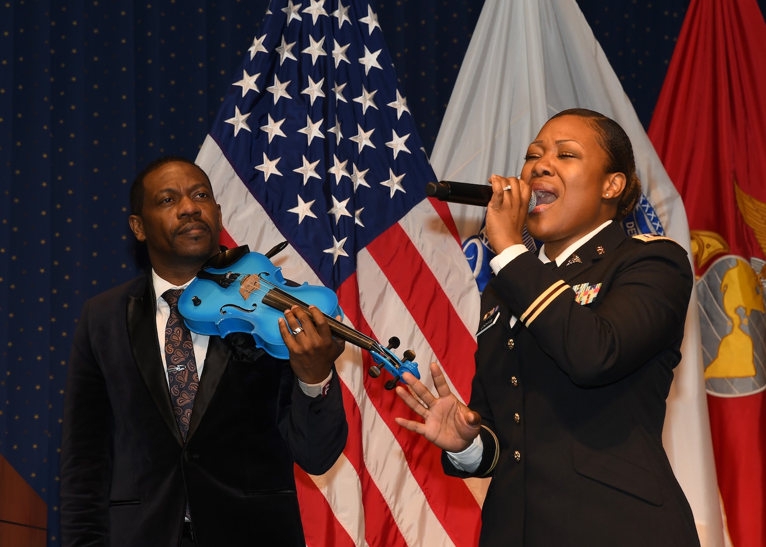 Man playing blue violin, woman singing, both on stage