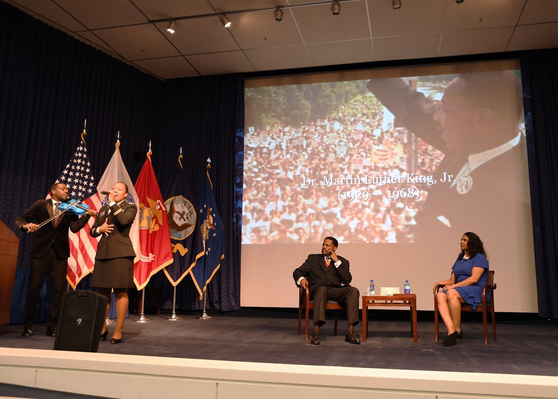 Man, woman seated on stage listening; man on violin and woman in Army uniform singing; MLK image in background