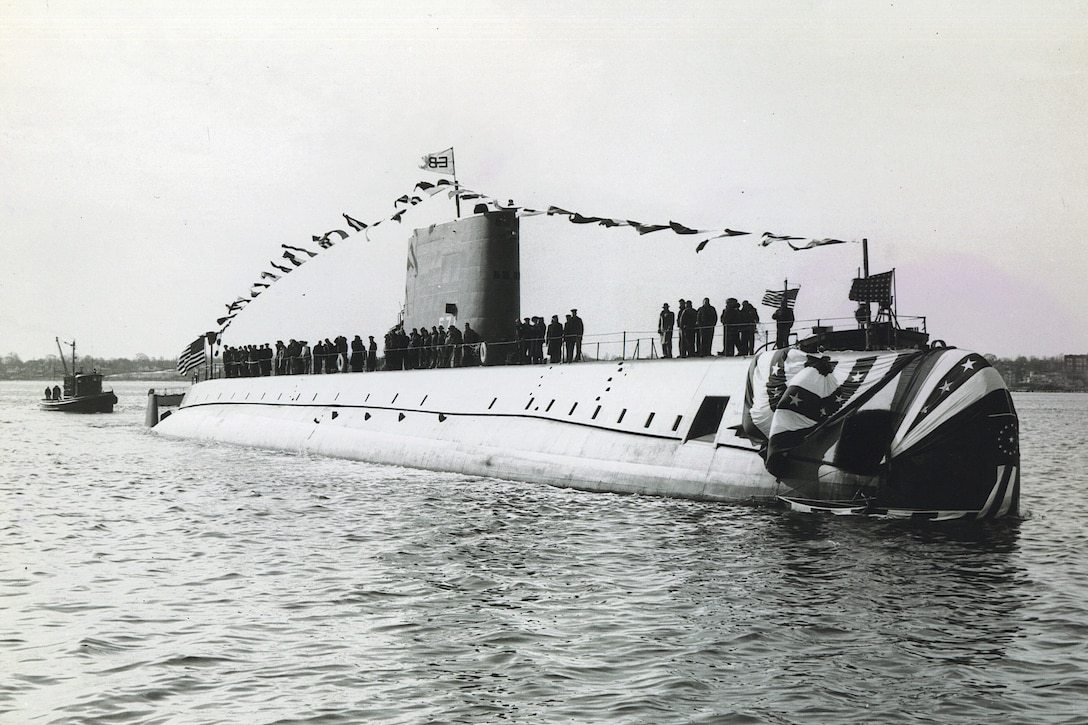 A submarine sits in the river as its submariners look ashore.