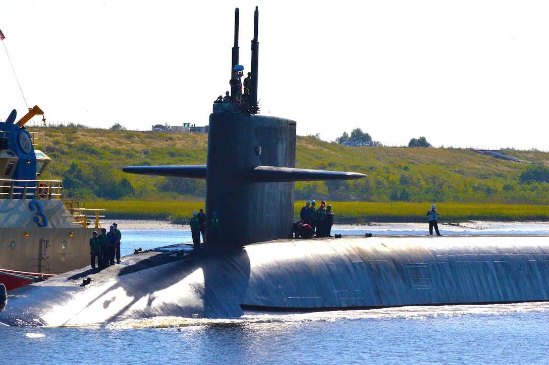 Sailors stand on and near the conning tower of a submarine near shore.