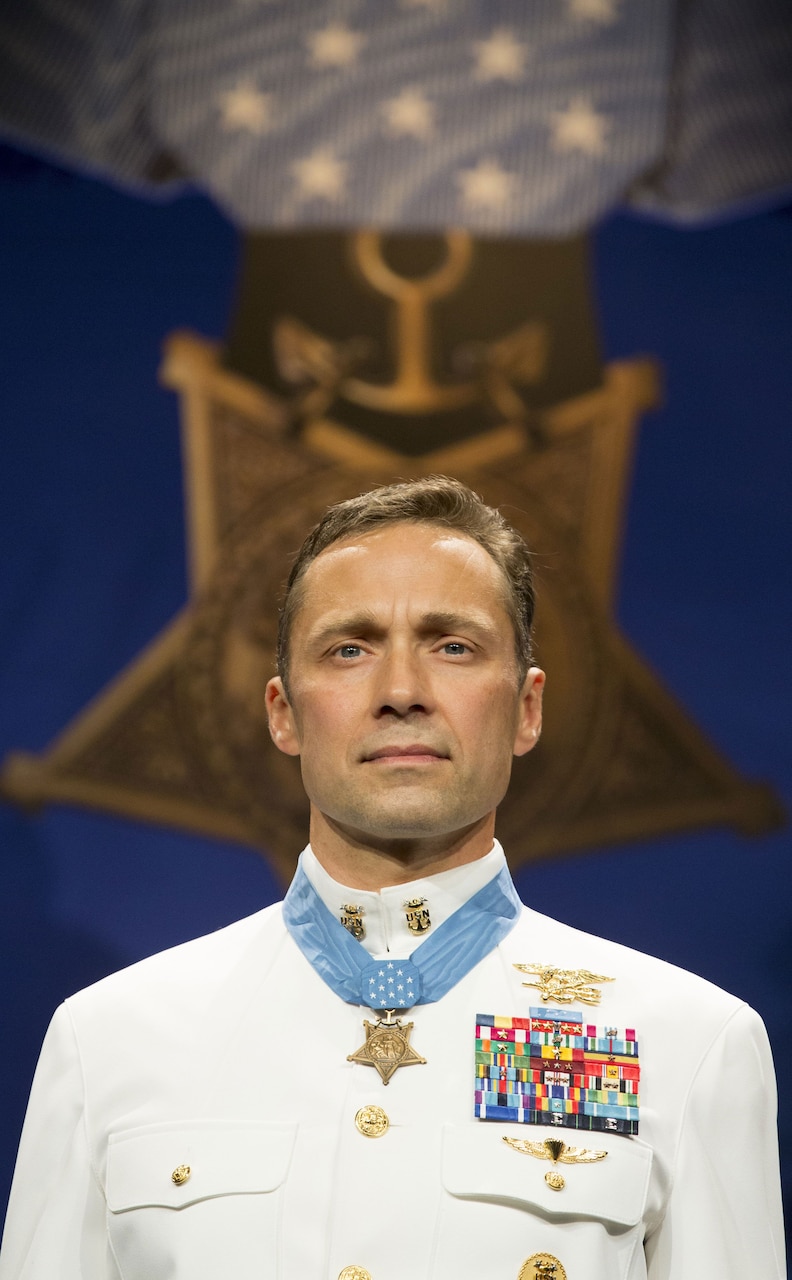 A Navy SEAL wearing the Medal of Honor stands in front of a large picture of the Medal of Honor.