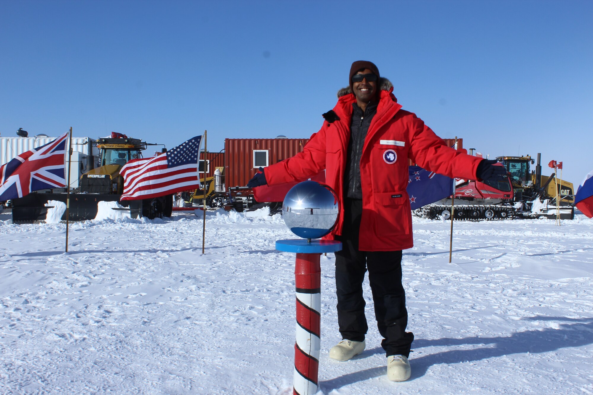 Capt. Michael Nayak, an AFRL scientist, at the Ceremonial South Pole with the South Pole Traverse (SPT) in the background. Several groups of trucks, bulldozers and Sno-Cats (tracked vehicles for snowy conditions) pull massive sleds with supplies from McMurdo Station to the South Pole Station. Each sled holds 20,000 gallons, or 140,000 pounds, of jet fuel. Every year, workers rebuild the thousand mile-road that supports this journey. (Courtesy photo)