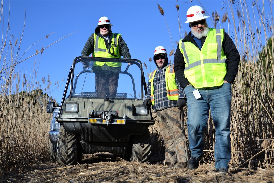 Three members of the USACE Water Resources Division, wearing yellow vests, stand near an all-terrain vehicle among phragmites at Joint Base Langley-Eustis