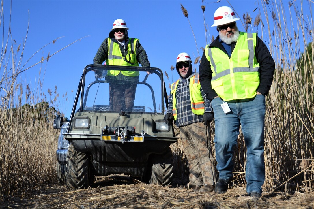 Three members of the USACE Water Resources Division, wearing yellow vests, stand near an all-terrain vehicle among phragmites at Joint Base Langley-Eustis