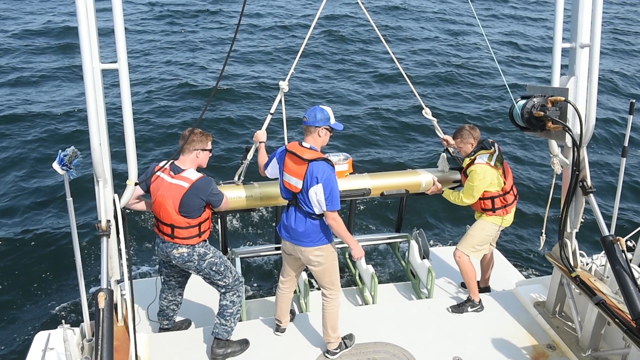 Three men on the back of a boat lower an autonomous underwater vehicle into water.