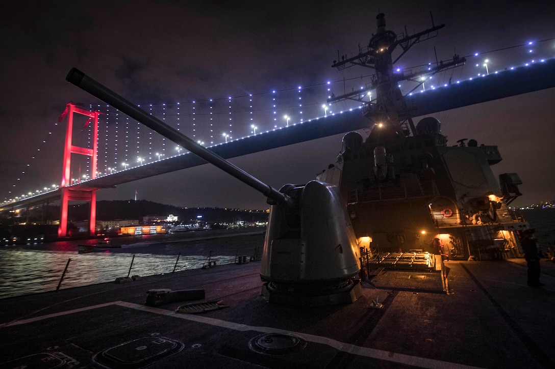 A military ship goes underneath a lit up bridge at night.