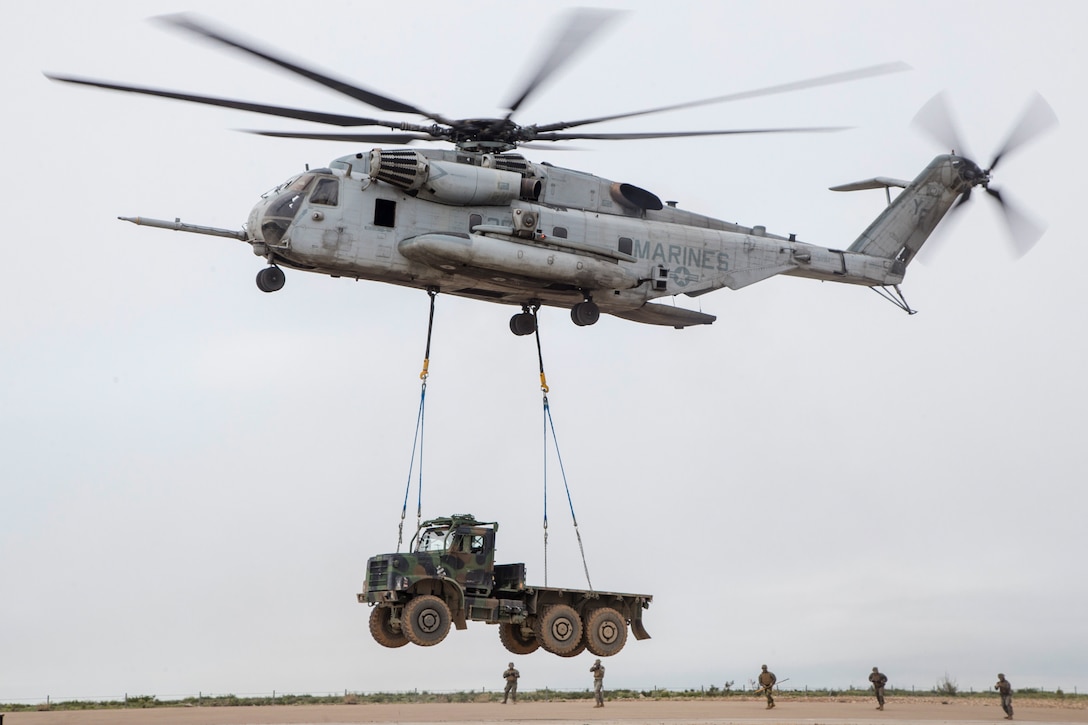 Five Marines stand underneath a helicopter lifting a truck.
