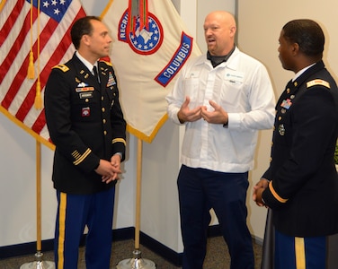 Three men talking. The two on the outside are wearing Army dress uniforms and the man in the middle is wear with top uniform from Honda and blue pants. Two flags in the background. The US flag and the Army Recruiting Flag.