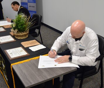 Two men sitting sign documents. One in white top from Honda and the other an Army soldier in dress blues. There is a plant in the middle of the table