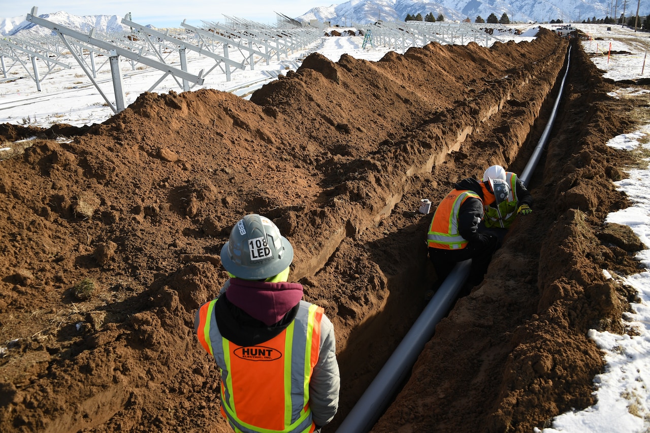 Workers in personal protective gear assemble a pipe inside a trench dug in a snowy field.