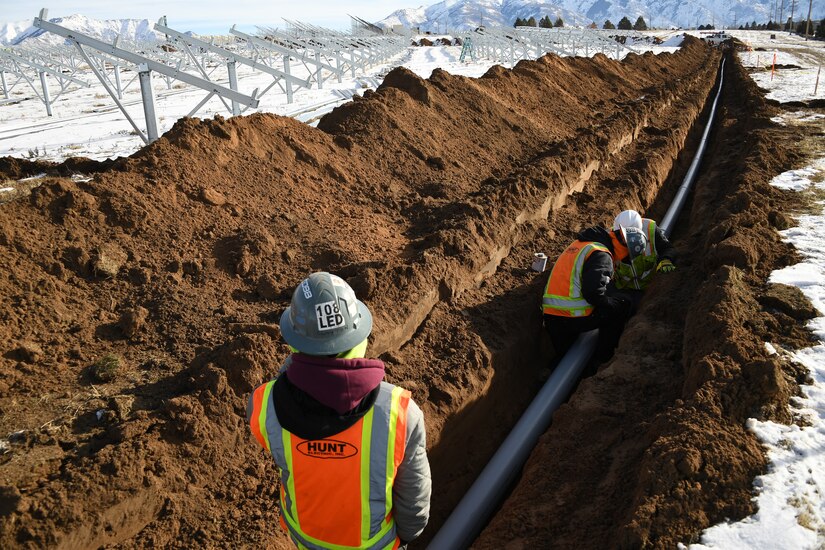 Workers in personal protective gear assemble a pipe inside a trench dug in a snowy field.