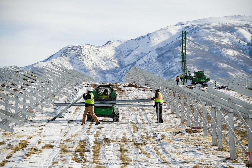 Workers in personal protective gear carry large metal brackets while building solar panels in a snowy field.