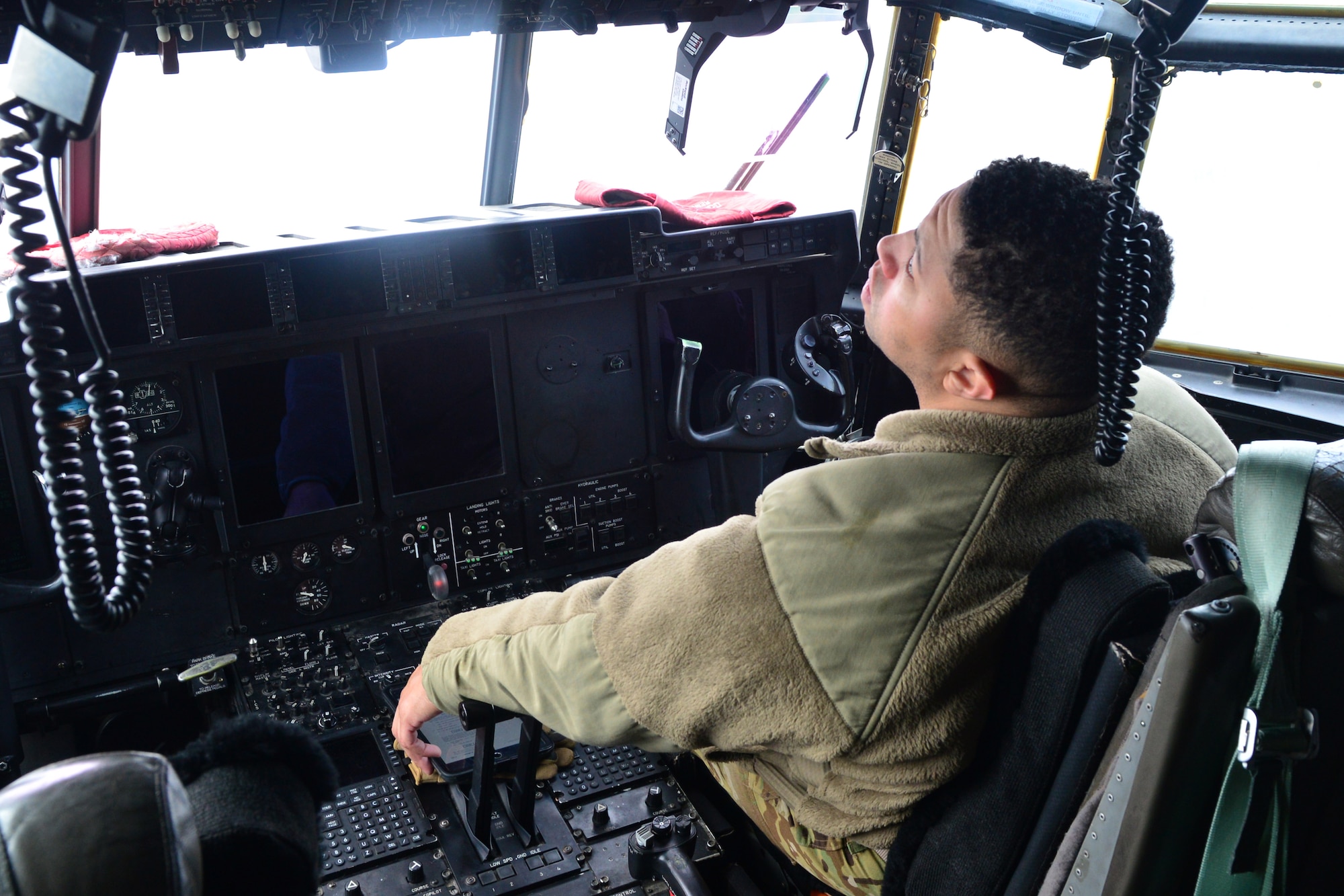 A man wearing the operational camouflage uniform fleece sits in the cockpit of a C-130J.