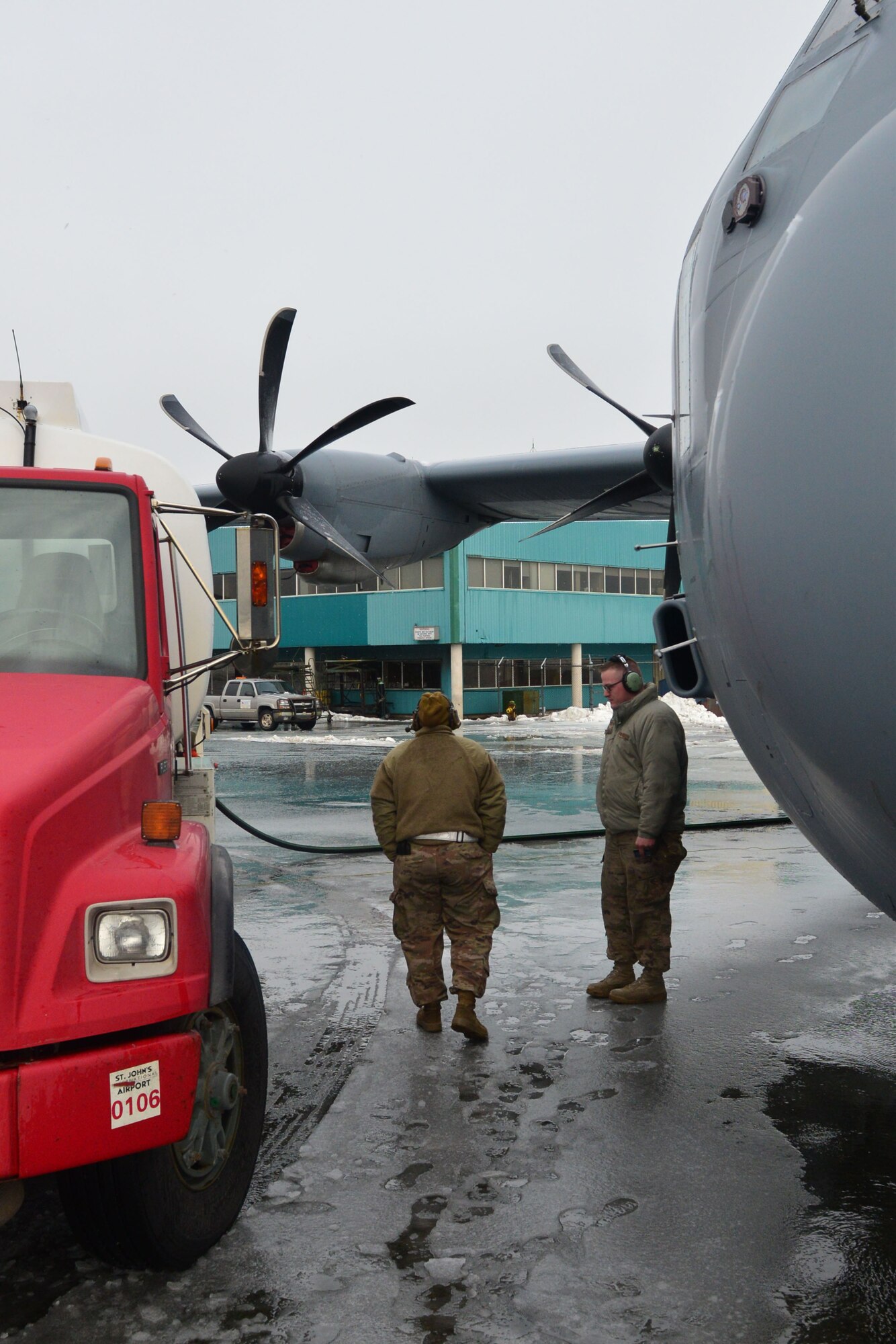 Two men stand between a truck and a C-130J.