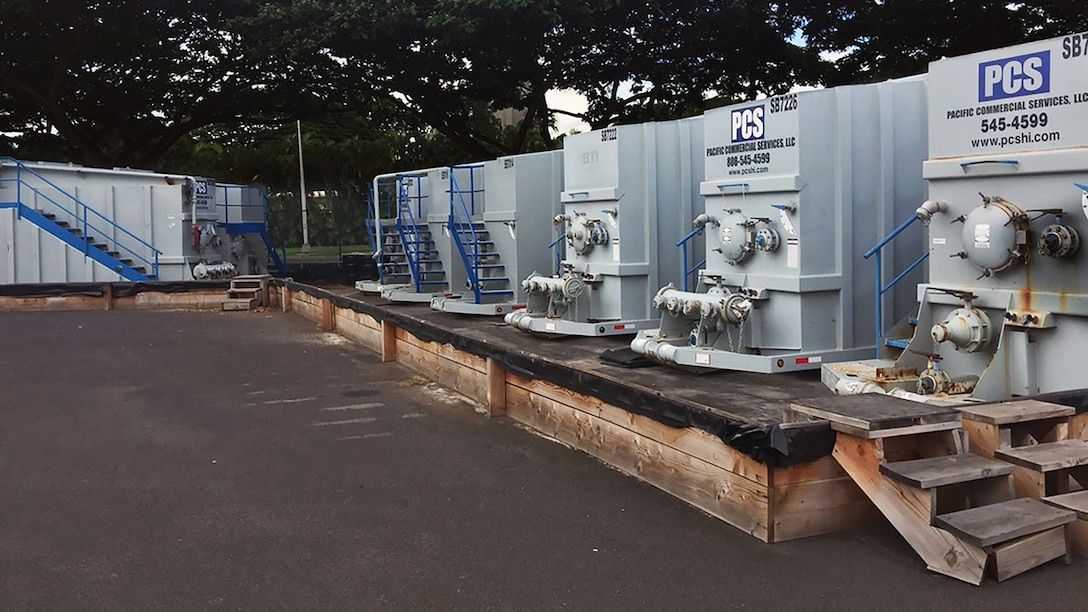 A row of at least six shipping container-sized fuel tanks lined up around a concrete pad.