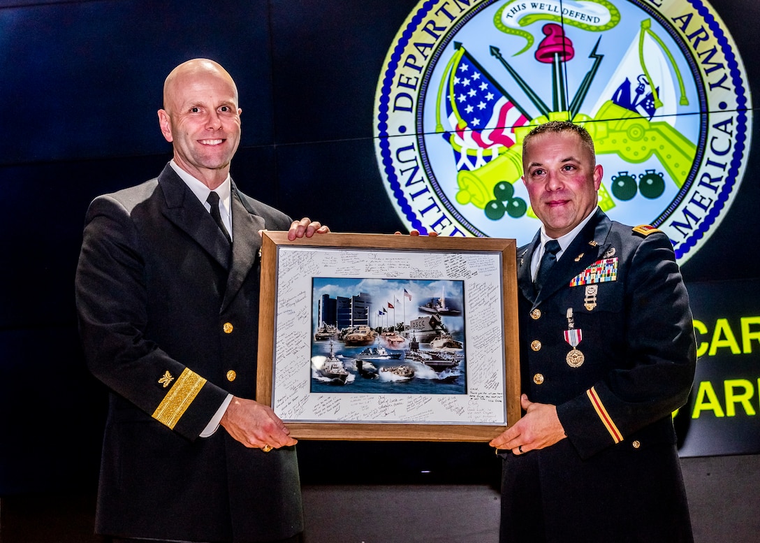 Two military officers stand on stage holding a frame with signatures around the edge and picture of the installation in the middle