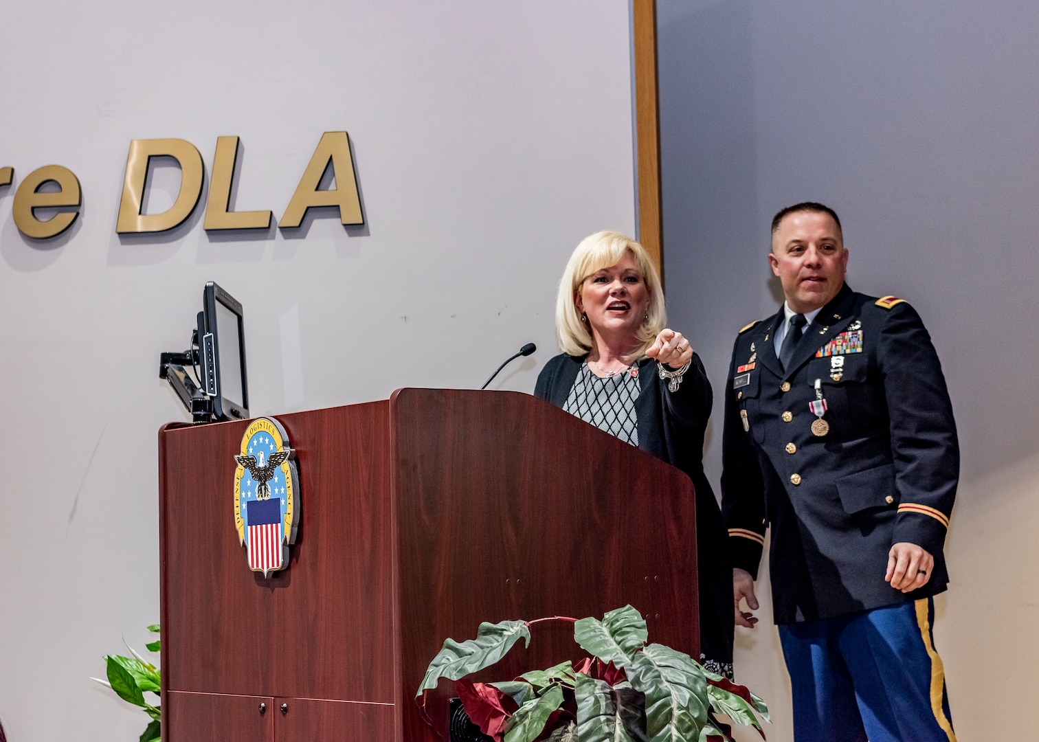 A woman points across the stage standing next to a military man, both behind a podium