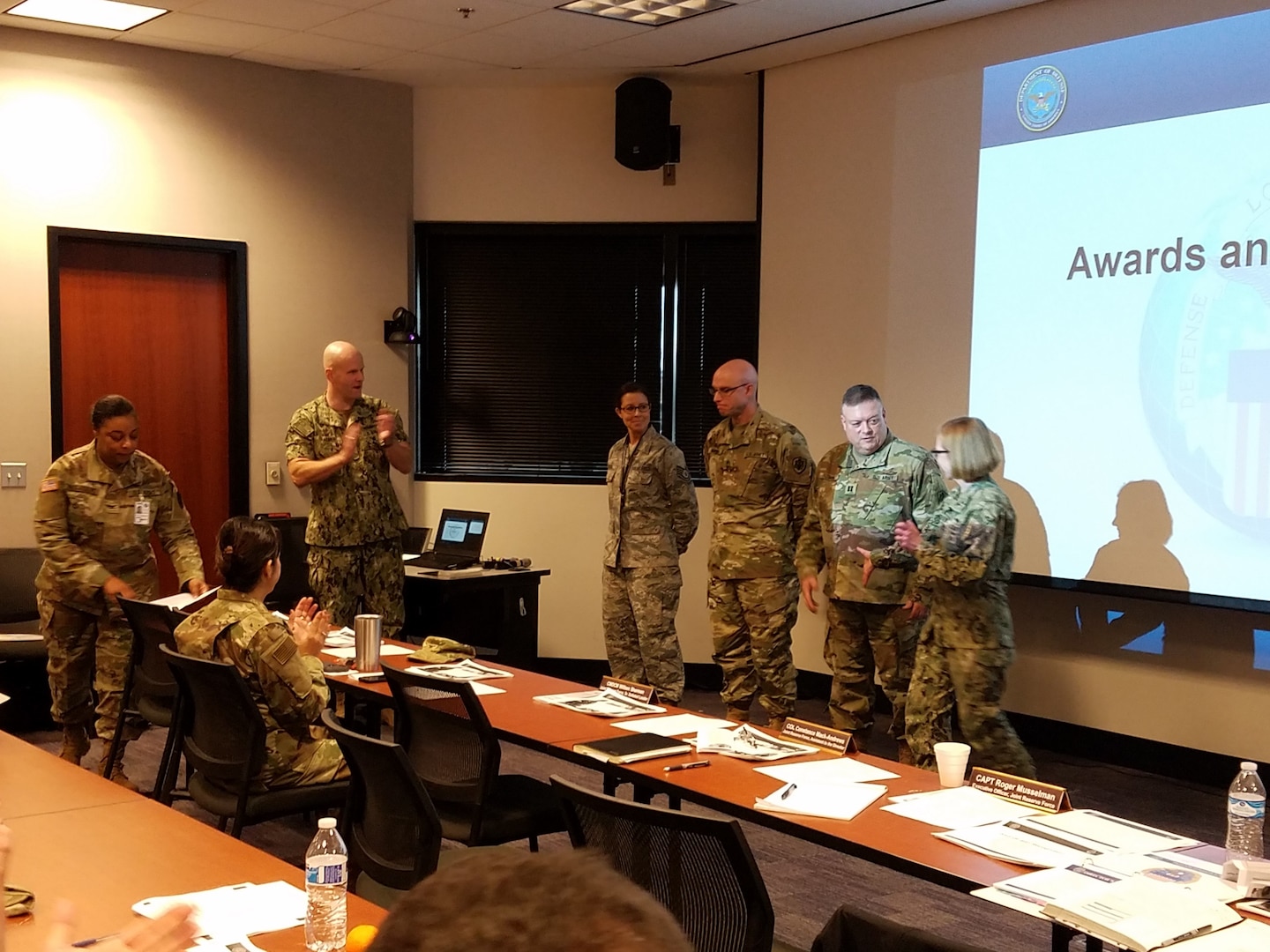 Four military members stand in front of a room of other military associates.