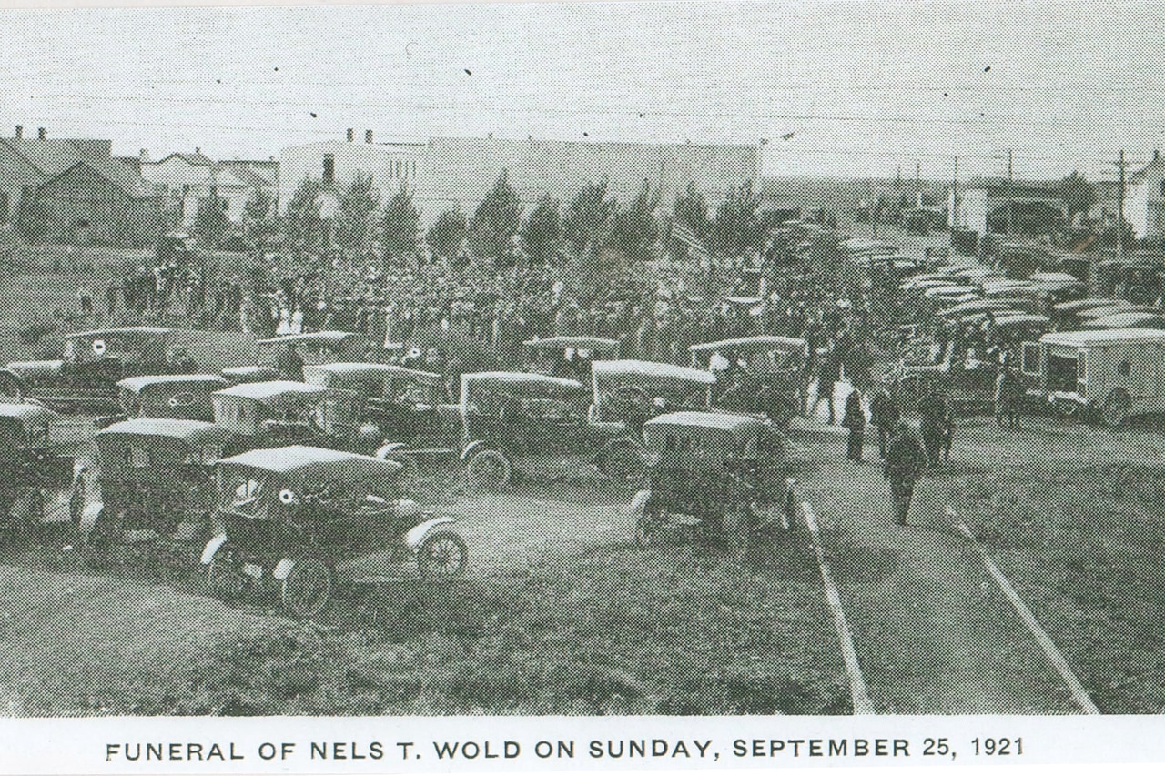 Several 1920’s-style cars surround a crowd at a funeral.