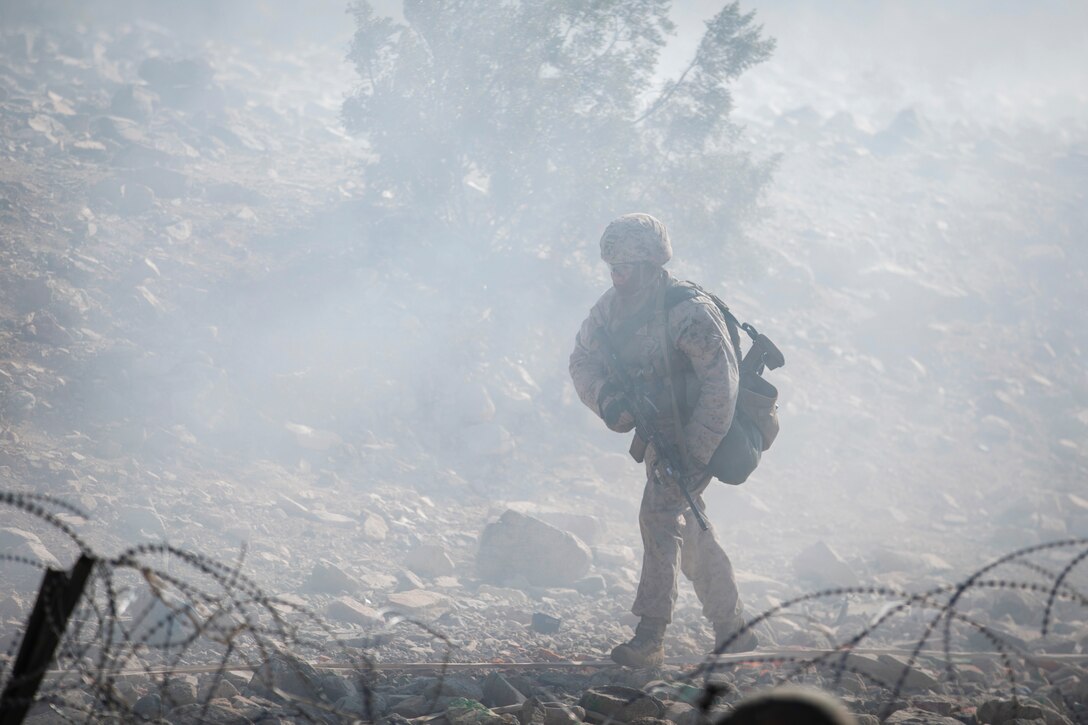 A Marine walks past barbed wire while surrounded by smoke.