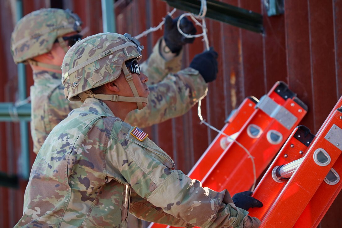 Two soldiers on ladders install concertina wire on a barrier structure.