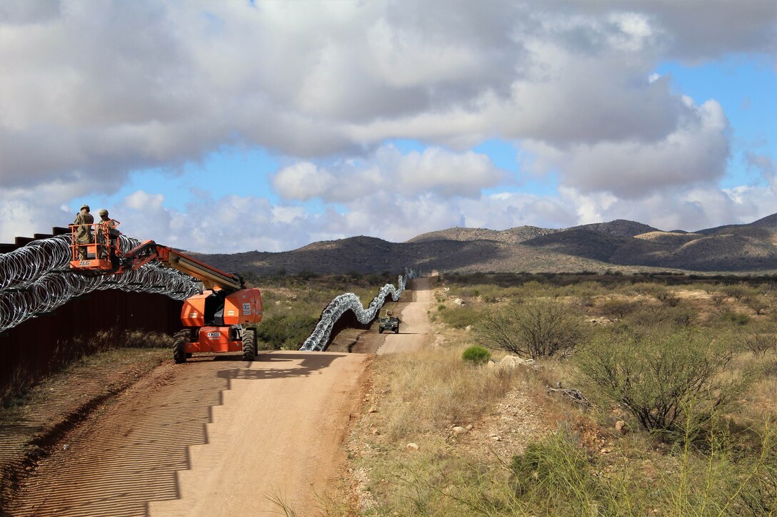 Soldiers work on placing wire along a long border structure over rolling hills.
