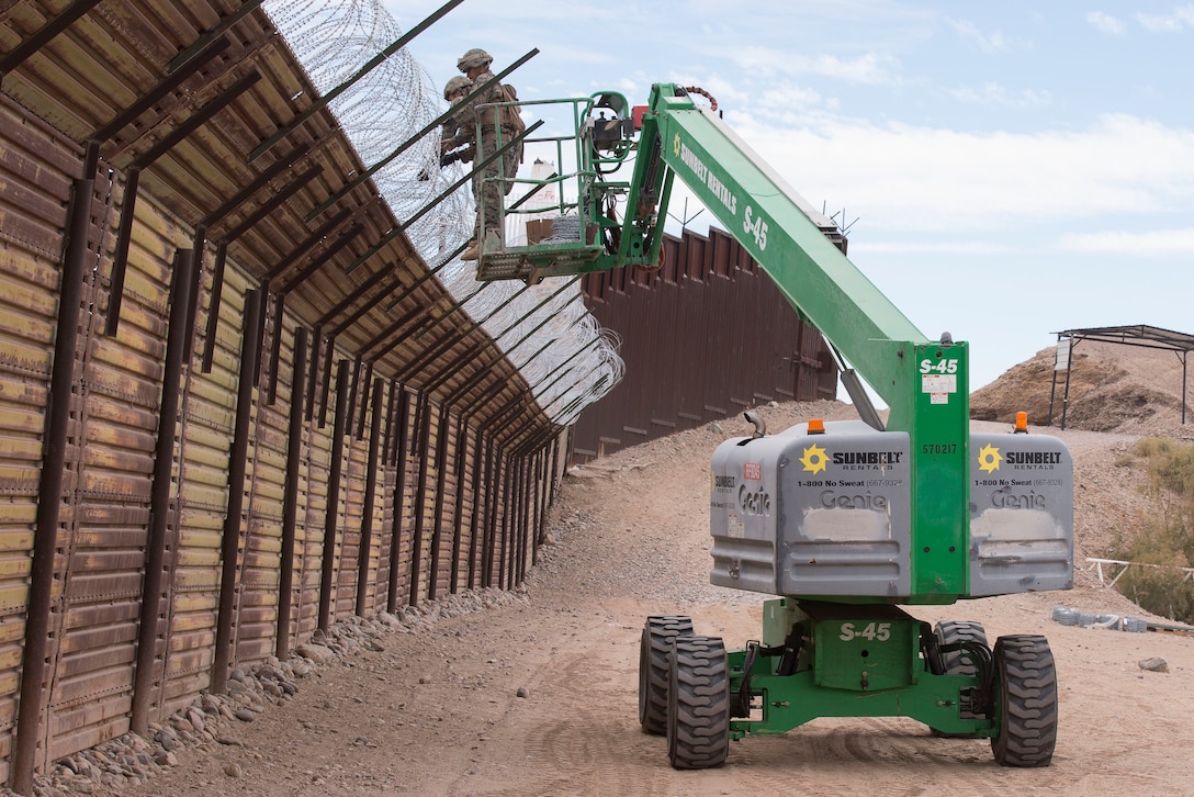 Marines standing on a platform attached to a construction vehicle secure wire atop a border barrier.