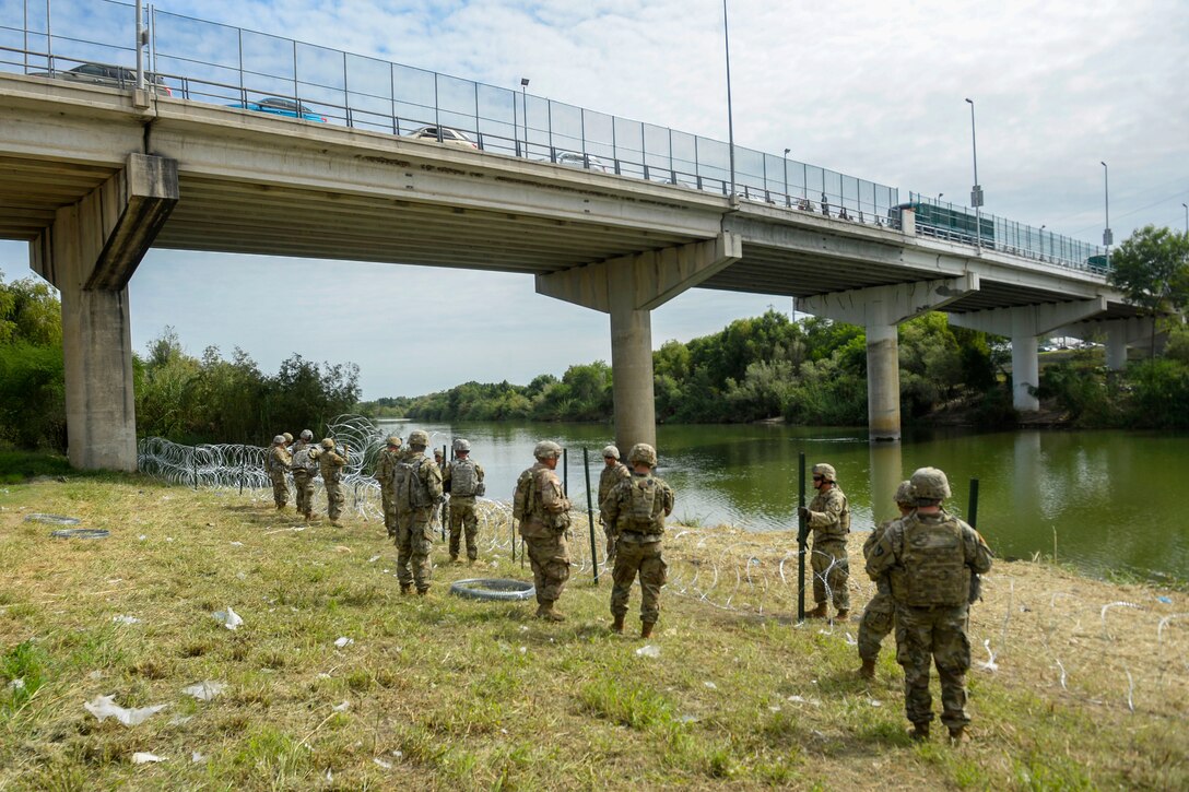 Soldiers place concertina wire on ground by a body of water below a bridge.