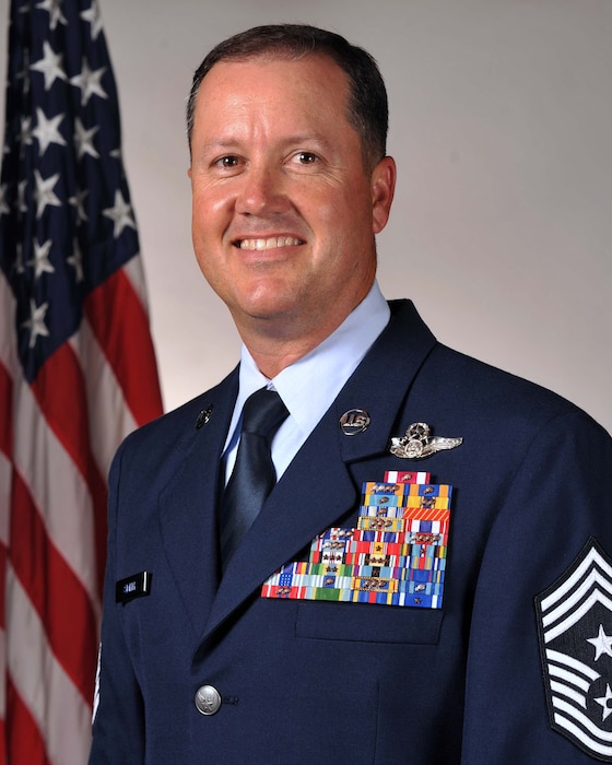White man in Air Force Blues uniform standing in front of flag.