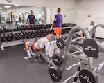 Weight room with a guy using free weights on a bench, another standing using free weights and another on a bench.