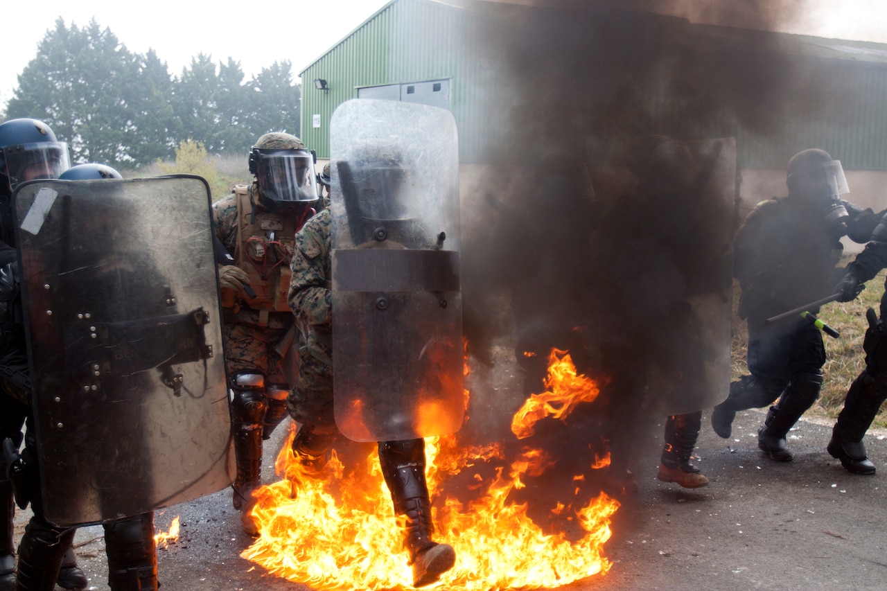 Marines and French gendarmes wearing riot control gear run through flames.