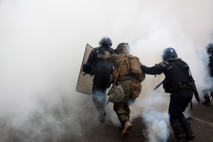 Marines and French gendarmerie in riot control gear run through a cloud of smoke.
