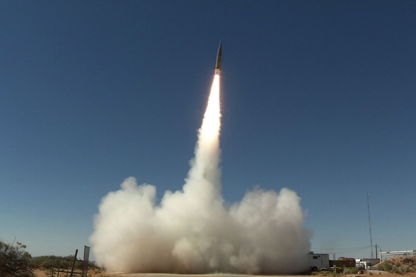 A missile launches skyward during a daytime test, leaving a trail of smoke and fire.