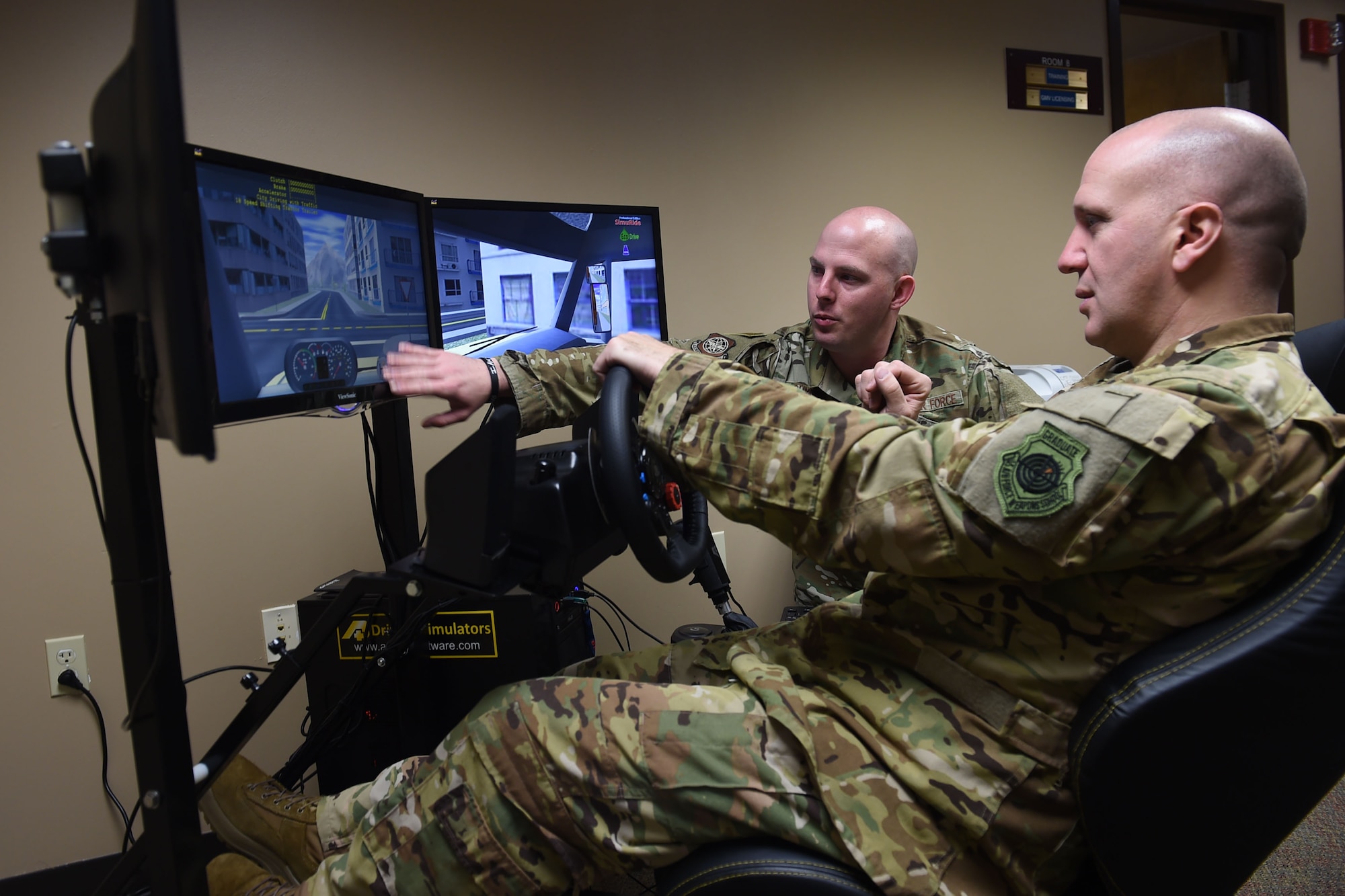 Two Airmen sit a three computer screens, one is sitting and using a steering wheel to control the drivng simulator on the screens.