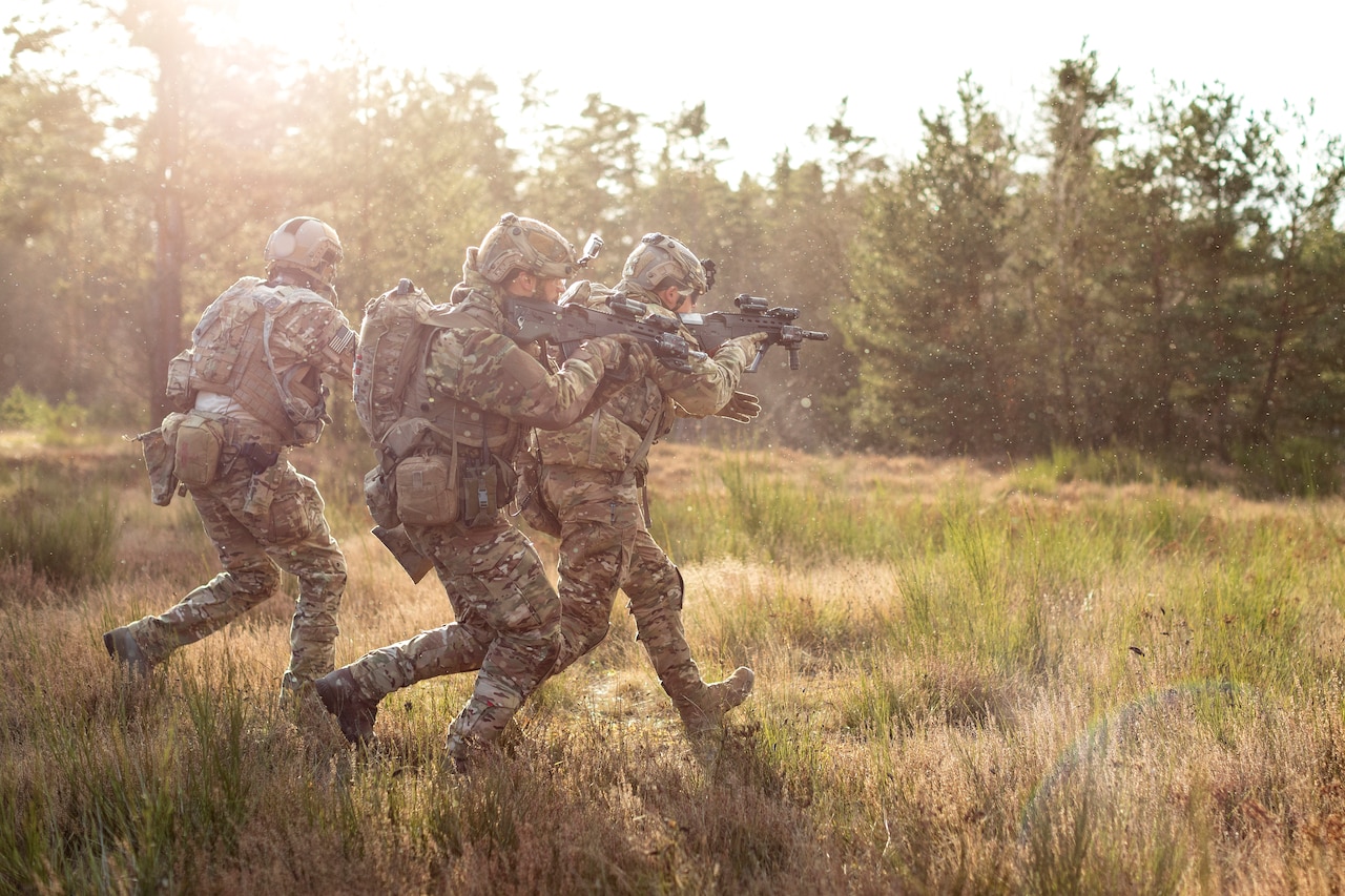 Three armed soldiers in camouflage move across a sunny field while pointing weapons.