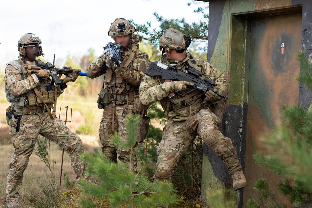 A soldier in camouflage kicks down a door as two others stand at the ready.